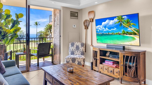 A spacious living room with a large TV, a wooden coffee table, a patterned armchair, and a view of the ocean through a sliding glass door. - Elegant Oceanfront Vistas (Kihei)
