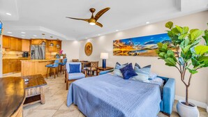 A modern hotel room with a bed, seating area, kitchenette, and a large painting of a sunset. - Elegant Oceanfront Vistas (Kihei)