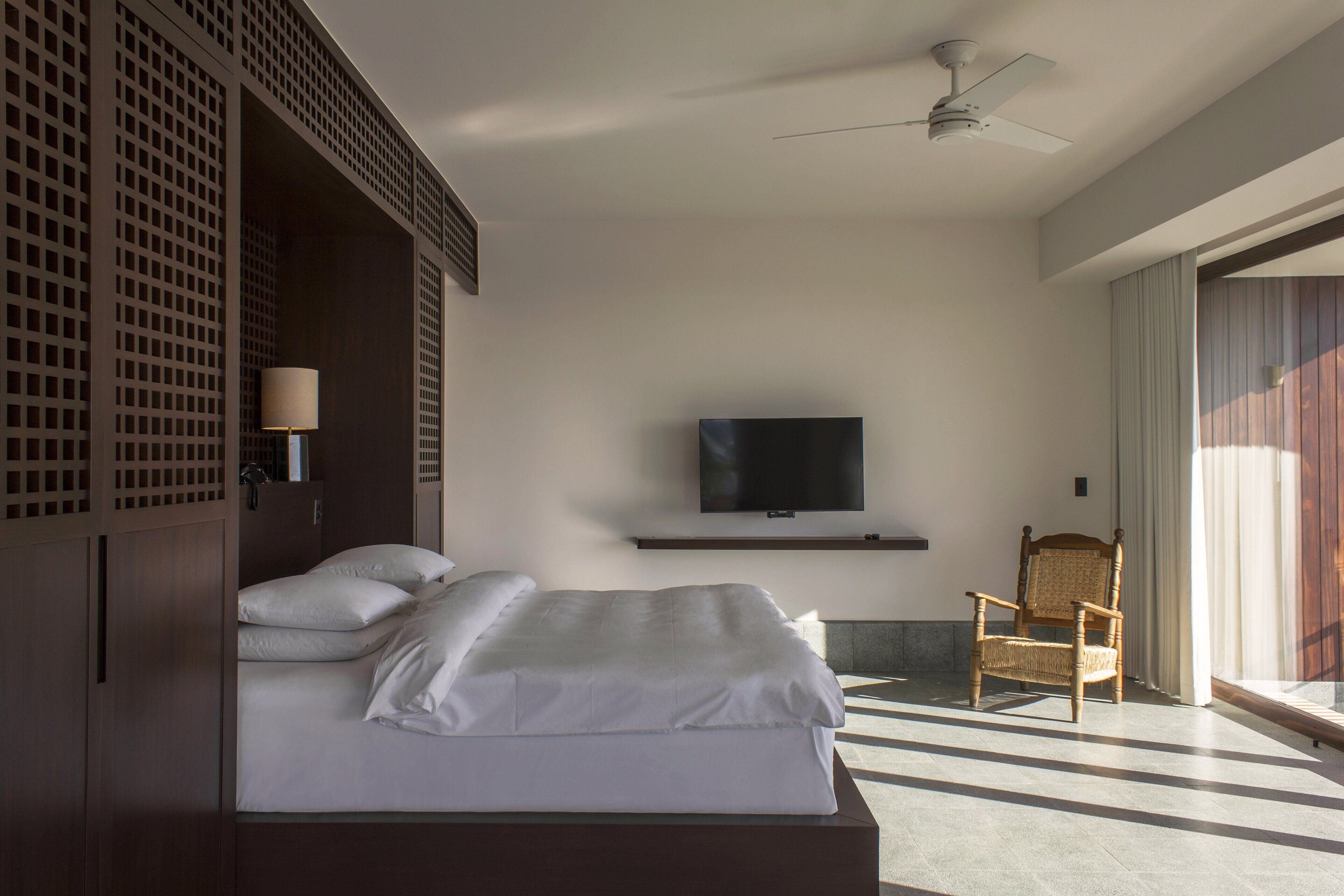 Una habitación de hotel moderna con una cama grande, un televisor montado en la pared, una silla y un ventilador de techo.