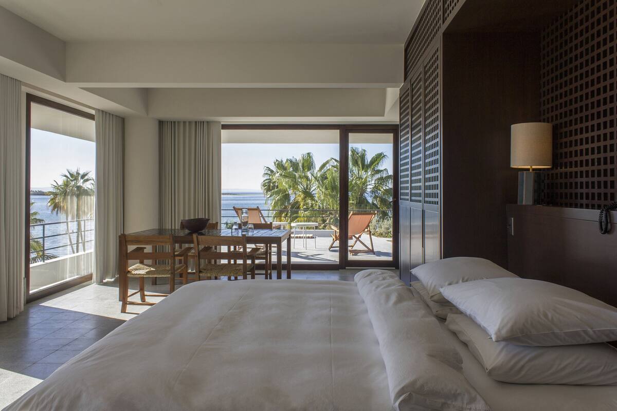 A hotel room with a large bed, a dining area with a table and chairs, and a view of the ocean and palm trees.