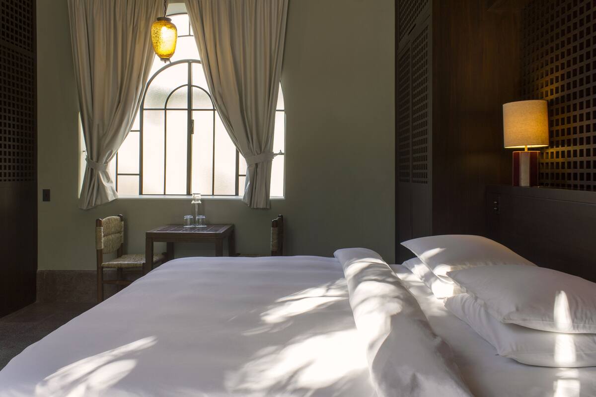 A hotel room with a bed, a chair, a small table with a glass, and a lamp.