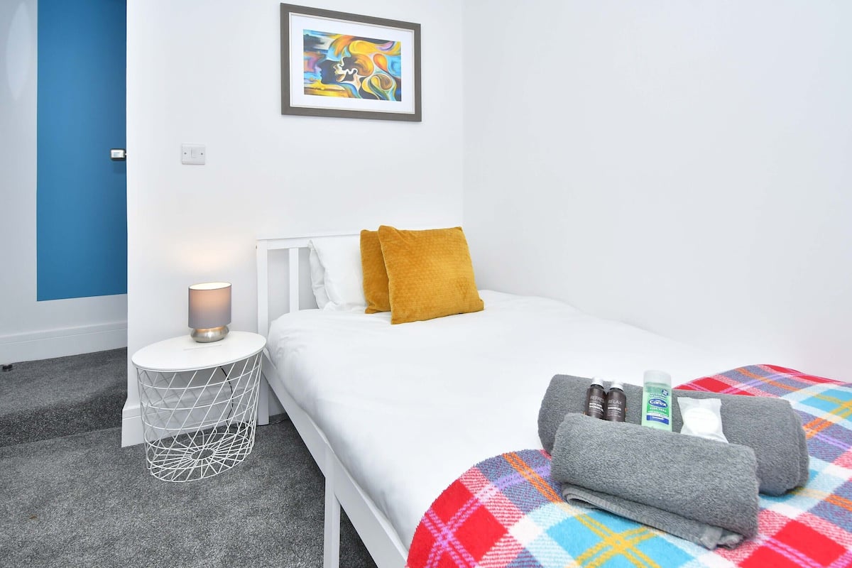 Un lit bien fait, avec une tête de lit blanche, un oreiller de couleur moutarde et une couverture colorée. Une petite table de chevet avec une lampe et un objet décoratif se trouve à côté du lit. Une œuvre d’art encadrée est accrochée au mur, au-dessus du lit.