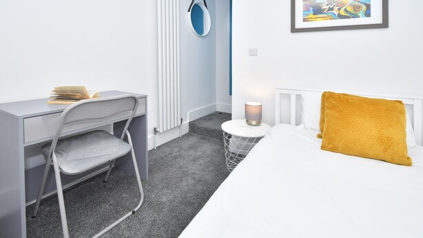 Ein kleines Hotelzimmer mit Bett, Schreibtisch mit Stuhl, rundem Tisch mit Lampe und einem gerahmten abstrakten Gemälde an der Wand.