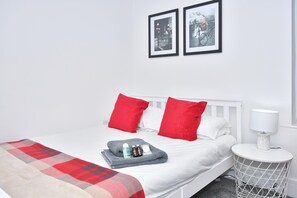 Un lit bien fait, avec des oreillers rouges et blancs, une couverture grise et une petite table de chevet sur laquelle se trouve une lampe.