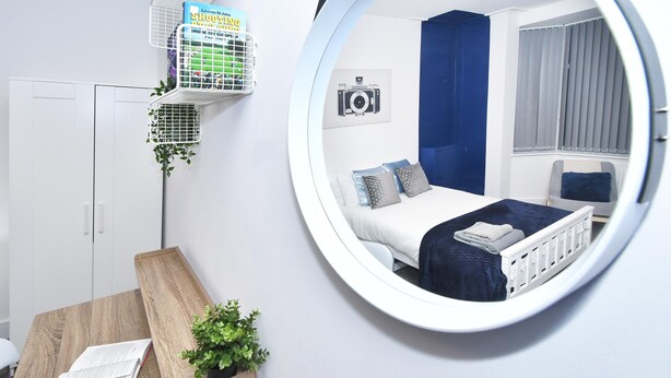 A bedroom with a round mirror, a bed with blue bedding, a white radiator, and a small shelf with a book and a plant. - Townhouse @ 42 Minshull New Road Crewe (Crewe)
