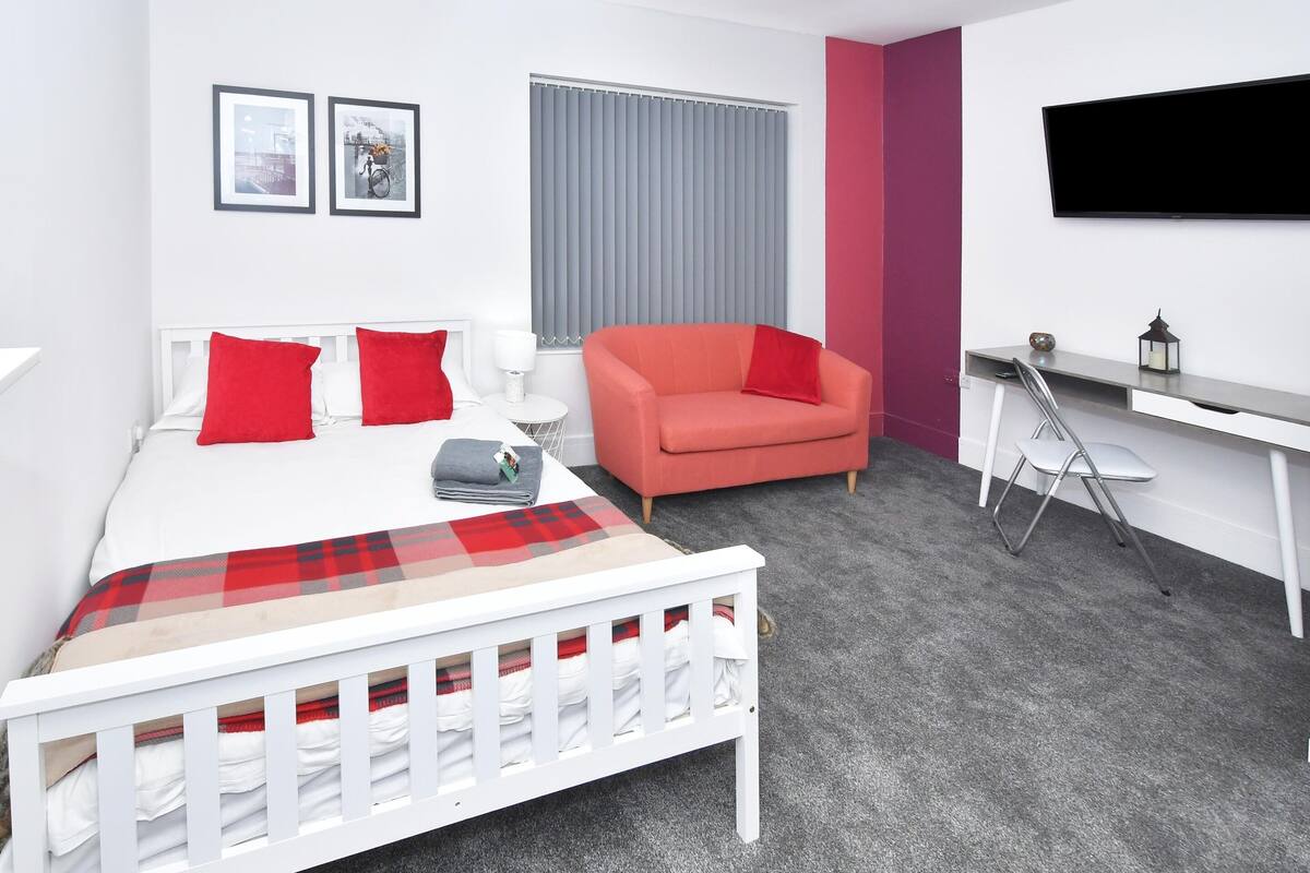 Une chambre d’hôtel moderne avec un lit blanc, des oreillers rouges, un canapé rouge, une moquette grise, un bureau blanc et un téléviseur fixé au mur.