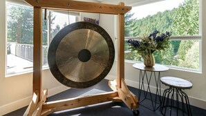 A wooden gong on a stand in a room with a view of trees through large windows. - Hidden Pond Gong Suite - Luxury , Pet Friendly, with Hot Tub and Private Beach  (Victoria)