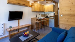A compact living space with a kitchenette, a TV mounted on the wall, a blue sofa, and a fruit bowl on a table. - Rond Point des Pistes (Val Claret) by Interhome (Tignes)