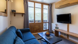 A modern living room with a sofa, a coffee table with fruit, a TV, and a large window with curtains. - Rond Point des Pistes (Val Claret) by Interhome (Tignes)