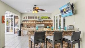 An outdoor kitchen with a stone island, bar stools, and a ceiling fan. - Private Beach, Heated Pool, Kayaks, & Pool Table - Villa Las Palmas - Roelens Vacations (Cape Coral)
