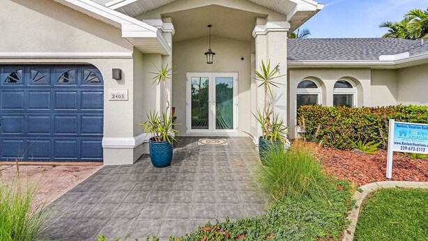 A two-story house with a blue garage door, a front entrance with a glass door, and a sign for Roelens Vacation Rentals. - Private Beach, Heated Pool, Kayaks, & Pool Table - Villa Las Palmas - Roelens Vacations (Cape Coral)