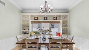 A dining room with a wooden table and chairs, a chandelier, and built-in shelving with decorative items. - Private Beach, Heated Pool, Kayaks, & Pool Table - Villa Las Palmas - Roelens Vacations (Cape Coral)