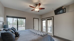 Un dormitorio con una cama, ventilador de techo, televisor montado en la pared y ventanas amplias.