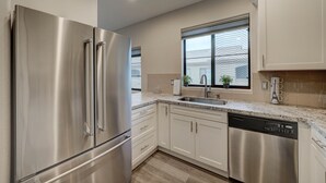 Una cocina moderna con electrodomésticos de acero inoxidable, armarios blancos, una encimera de granito y una ventana con vistas a edificios.