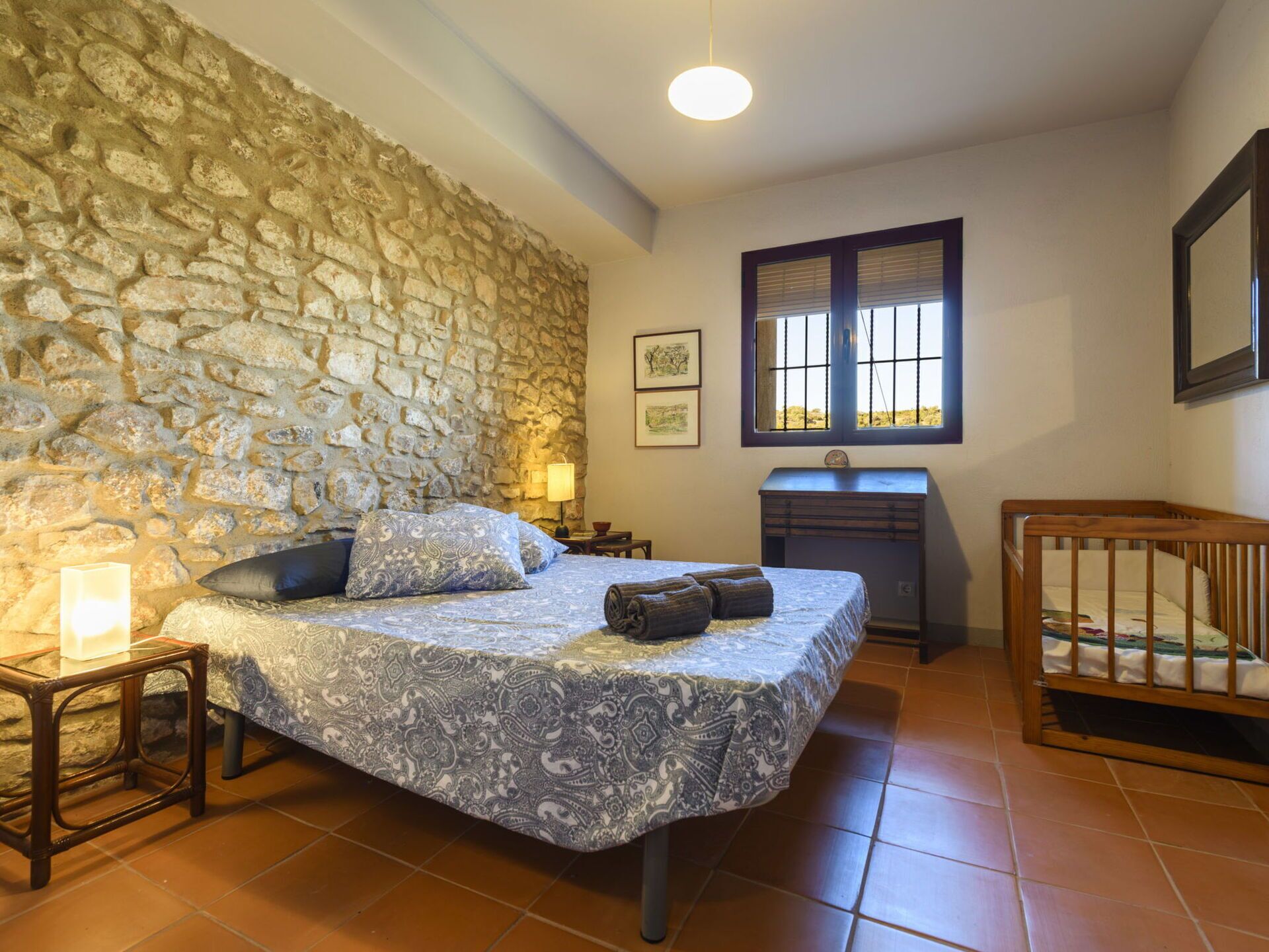 A bedroom with a stone wall, a bed with patterned bedding, a nightstand, a desk, a crib, and a window with blinds.