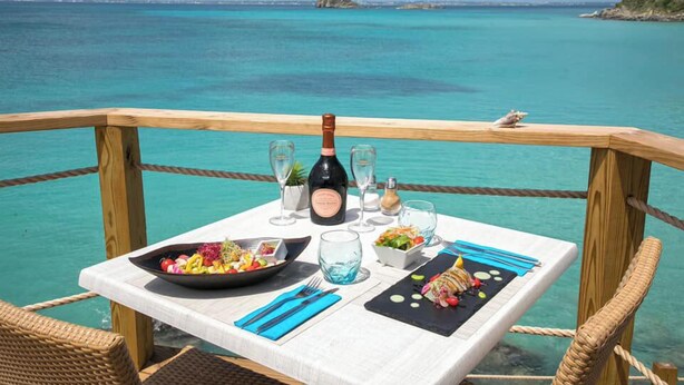 Mesa para dos con botella de vino, copas y comida, con vista al mar turquesa.