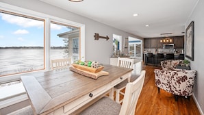 A dining area with a wooden table and chairs, a view of the water, and a kitchen in the background. - Beautiful Sunrise Views of Hall Lake! (Fairmont)