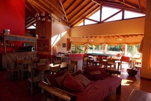 A spacious room with wooden furniture, a bar area, and large windows offering a view of the outdoors. - Hostel Inn Calafate (El Calafate)