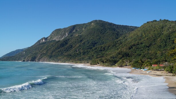 A beach with clear turquoise waters, a sandy shore, and a mountain in the background.