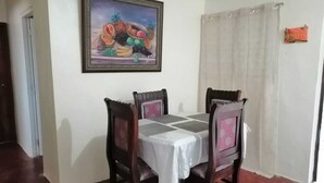 A dining area with a table set for four, surrounded by wooden chairs with patterned cushions. A framed painting of fruits hangs on the wall above the table.