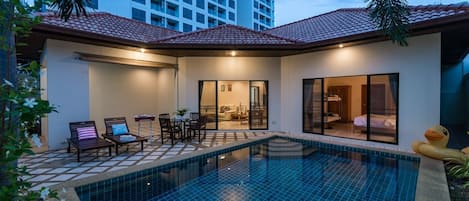 A modern pool area with a tiled floor, a seating area with chairs and a table, and a glass-enclosed room with a bed.