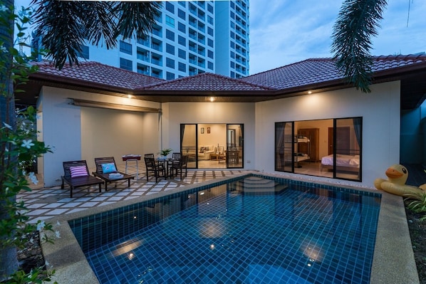 A modern pool area with a tiled floor, a seating area with chairs and a table, and a glass-enclosed room with a bed.