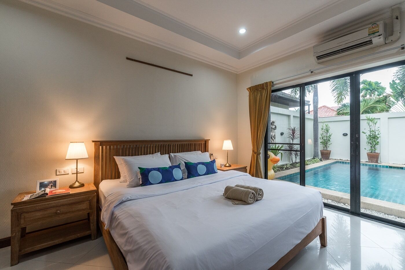 Habitación de hotel con una cama grande, dos mesitas de noche con lámparas, vista a una piscina y un equipo de aire acondicionado.