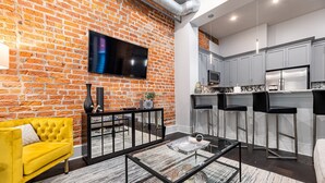 A modern living room with a brick wall, a yellow sofa, a glass coffee table, and a kitchen area with grey cabinets and a stainless steel refrigerator. - ⚜️ Perfect Downtown 1 Bedroom plus Queen Sleeper Sofa & Private Terrace ⚜️ - The Livingston (New Orleans)