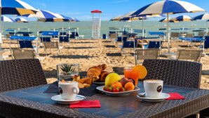 Een tafel aan het strand met ontbijtartikelen, een glas sinaasappelsap en uitzicht op het strand met parasols en stoelen.