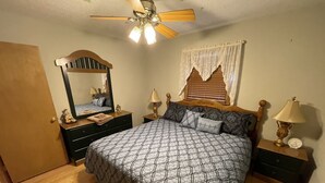Un dormitorio con cama, mesita de noche, cómoda, ventilador de techo y ventana con cortinas.