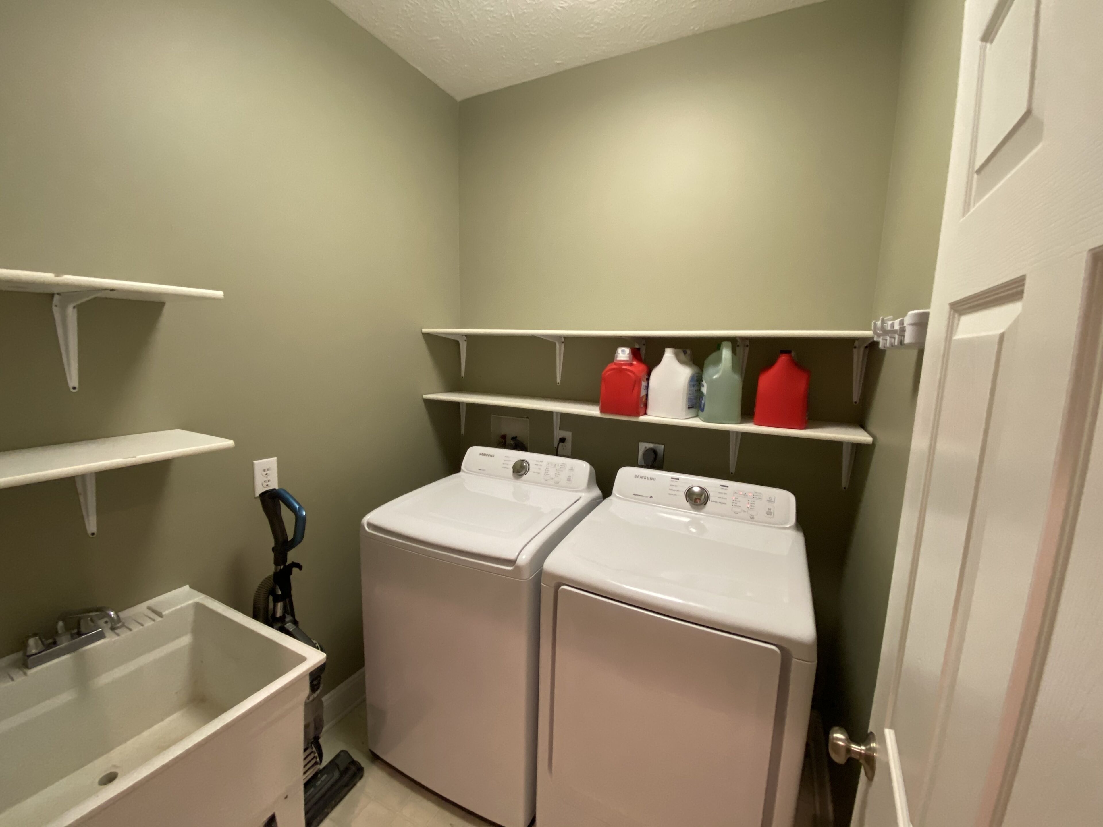 Un cuarto de lavandería con dos lavadoras frontales blancas, repisas con botellas de detergente y un tendedero de metal.