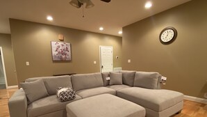 Ein Wohnzimmer mit Ecksofa, Hocker, Holzboden, einer Uhr an der Wand und einer Tür im Hintergrund.