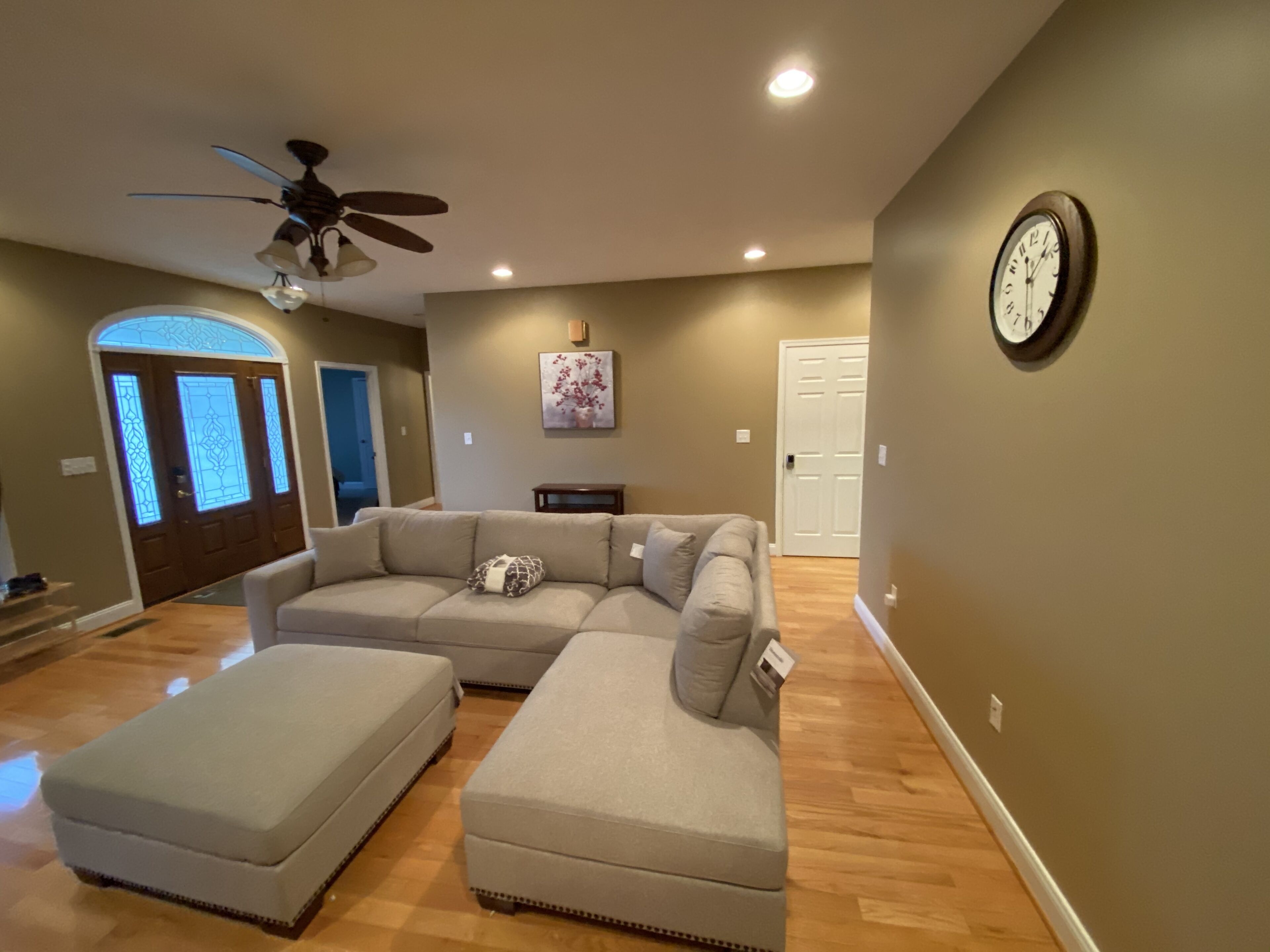 A spacious living room with a sectional sofa, ottoman, ceiling fan, recessed lighting, and a clock on the wall.