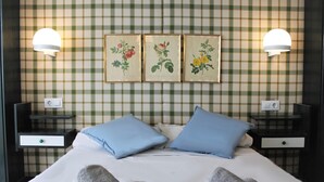 Een net opgemaakt bed met blauwe kussens, twee nachtkastjes met lampen, en behang met ruitpatroon.