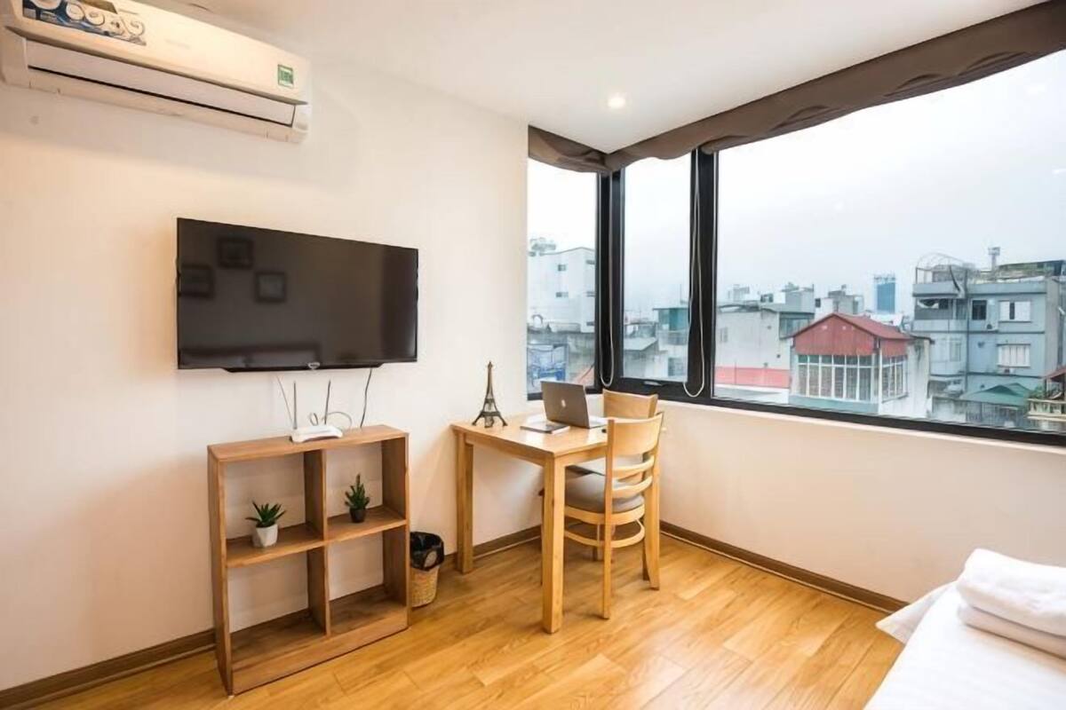 A room with a TV, wooden desk, chair, and a large window with a city view.