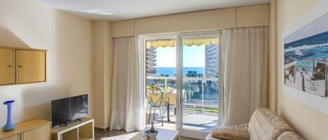 A living room with a television, a sofa, a table with a bottle, and a view of the ocean through a large window.