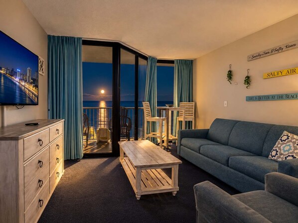 A living room with a sofa, a coffee table, and a TV on a stand. There is a balcony with a view of the city and the sea. - Oceanfront Luxe | Sand Dunes Eleven Twenty-Nine (Myrtle Beach)