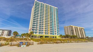 A tall hotel with multiple balconies, a sandy beach in the foreground, and a clear blue sky. - Towers On the Grove 724 (North Myrtle Beach)