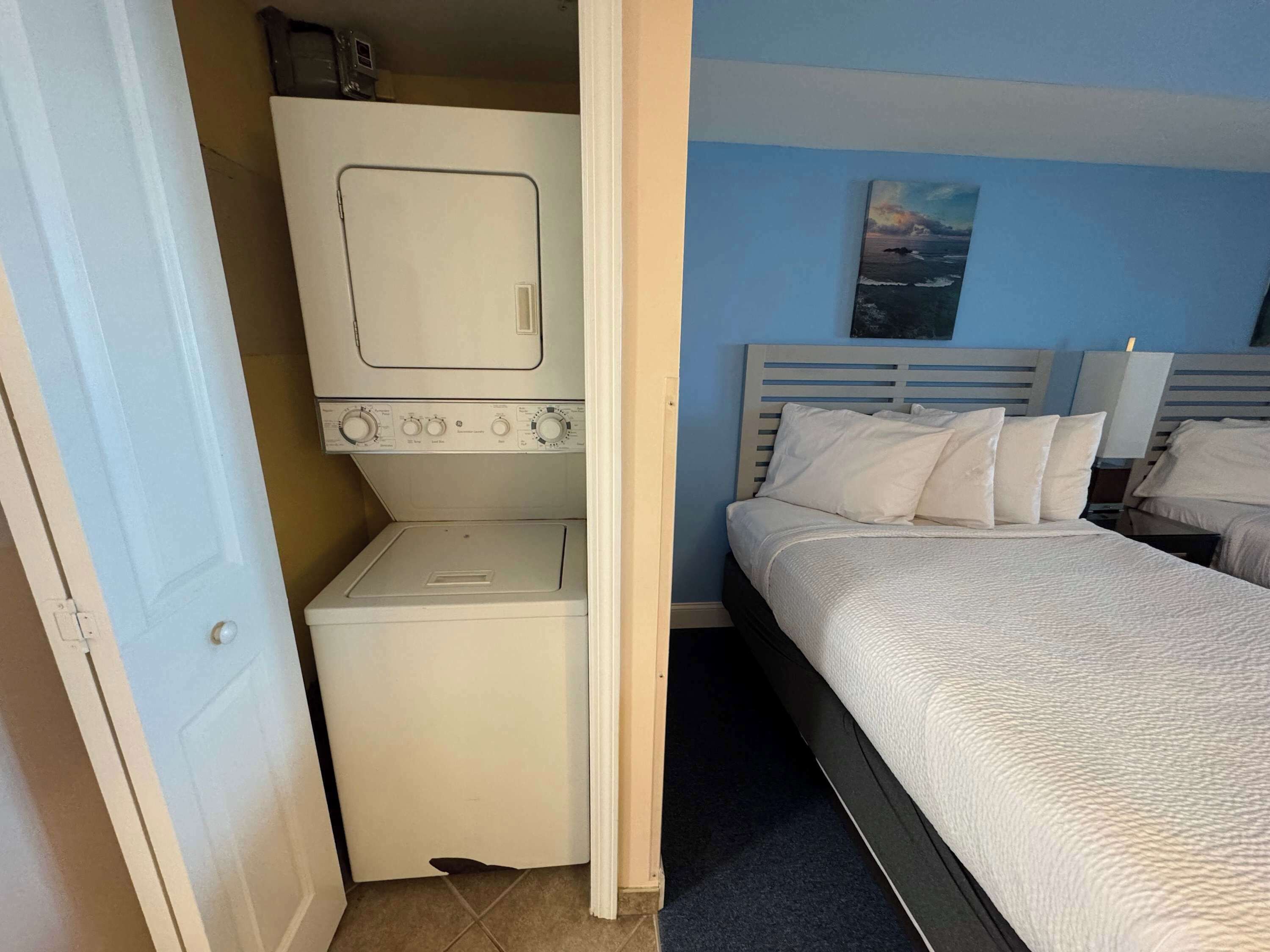 A room with a bed, a washing machine, and a painting on the wall.