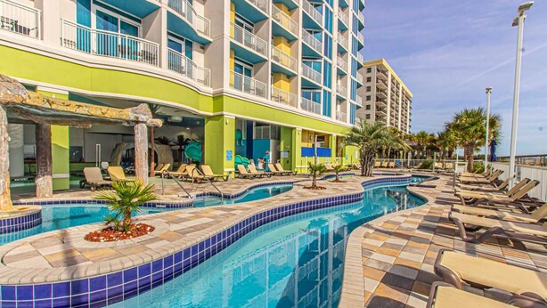 A hotel pool area with lounge chairs, a swimming pool, and a multi-story building with balconies. - Towers On the Grove 724 (North Myrtle Beach)