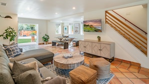 A spacious living area with a sofa, coffee table, and ottoman, a dining table with chairs, a cabinet, and a staircase. - Secluded Luxury Haven | Hot Tub, Billiards, Firepit & Spa Ambiance near Beach (Montecito)