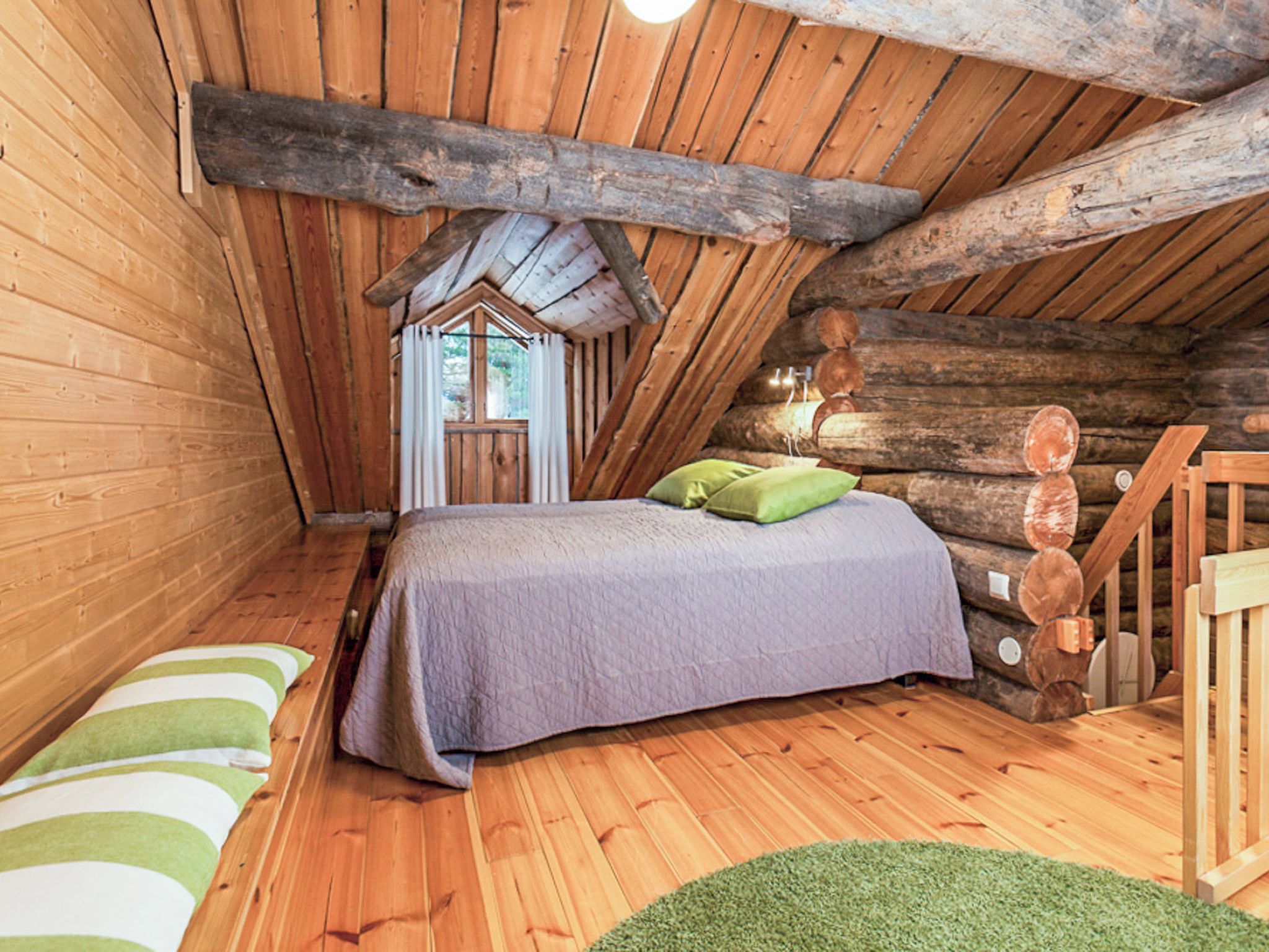 Un dormitorio acogedor en el ático, con techo inclinado, paredes de madera, una cama con colcha morada y una ventana pequeña.