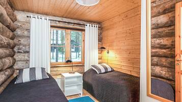A log cabin room with two beds, a window with curtains, a lamp, and a view of trees outside.