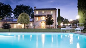 Une piscine avec des chaises longues, un bâtiment en briques surmonté d’un toit en tuiles et une pelouse bien entretenue.