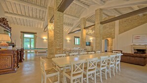 Une salle à manger spacieuse avec de hauts plafonds et des poutres apparentes, comprenant une longue table et des chaises blanches.