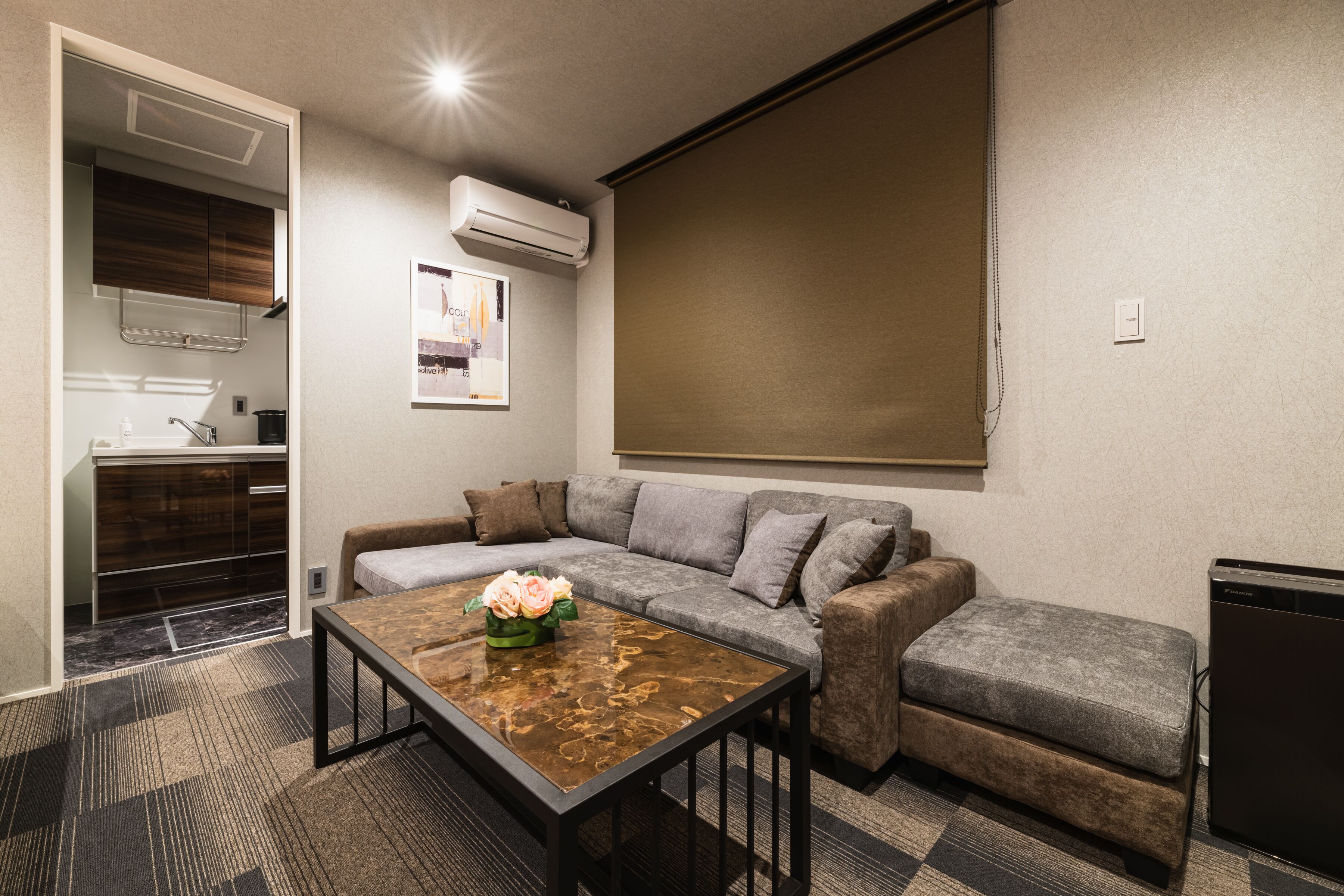 Un salon moderne avec un canapé, une table basse et un petit pouf. On y trouve également un coin cuisine équipé d’un évier et de placards, ainsi qu’un appareil de climatisation fixé au mur.
