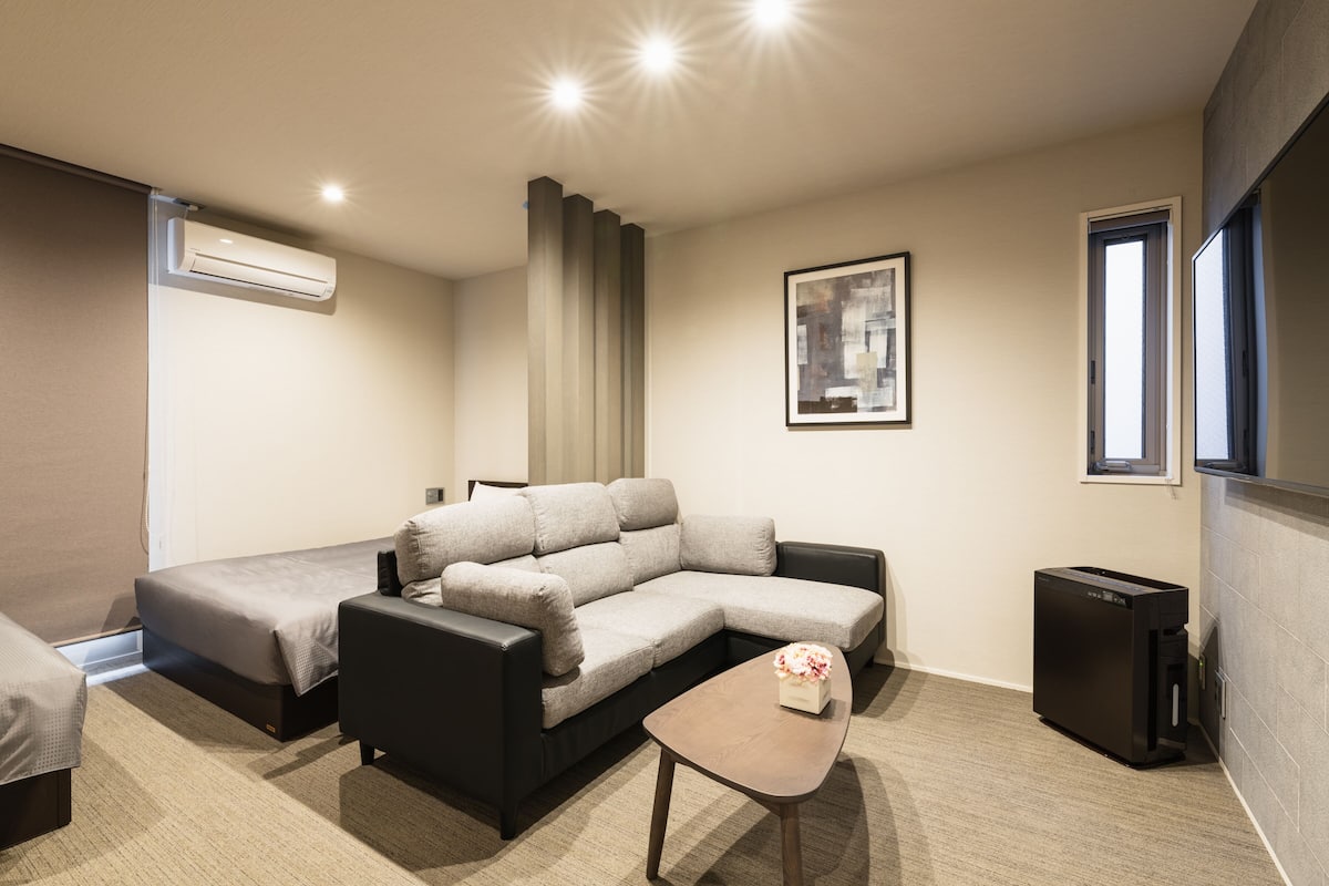 A modern hotel room with a sectional sofa, a small table, a trash bin, and a framed picture on the wall.