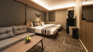 A hotel room with a sofa, two beds with decorative headboards, a small table with flowers, a trash bin, and a television on a stand. - GRAND BASE Kagoshima Tenmonkan (Kagoshima)