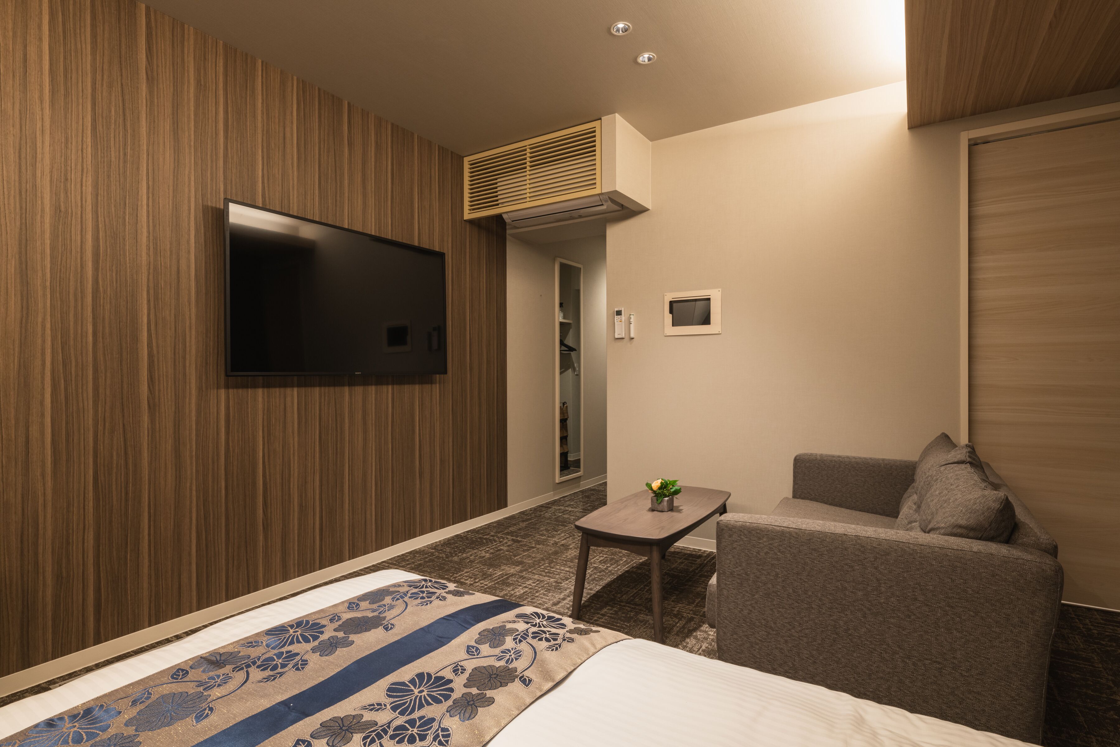 A hotel room with a bed, a TV mounted on the wall, a small table with a plant, a sofa, and a wooden panel wall.