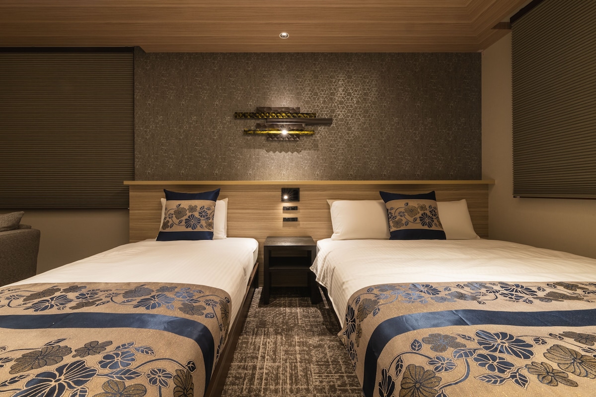 A hotel room with two beds, a nightstand, a wall-mounted light fixture, and a patterned rug.